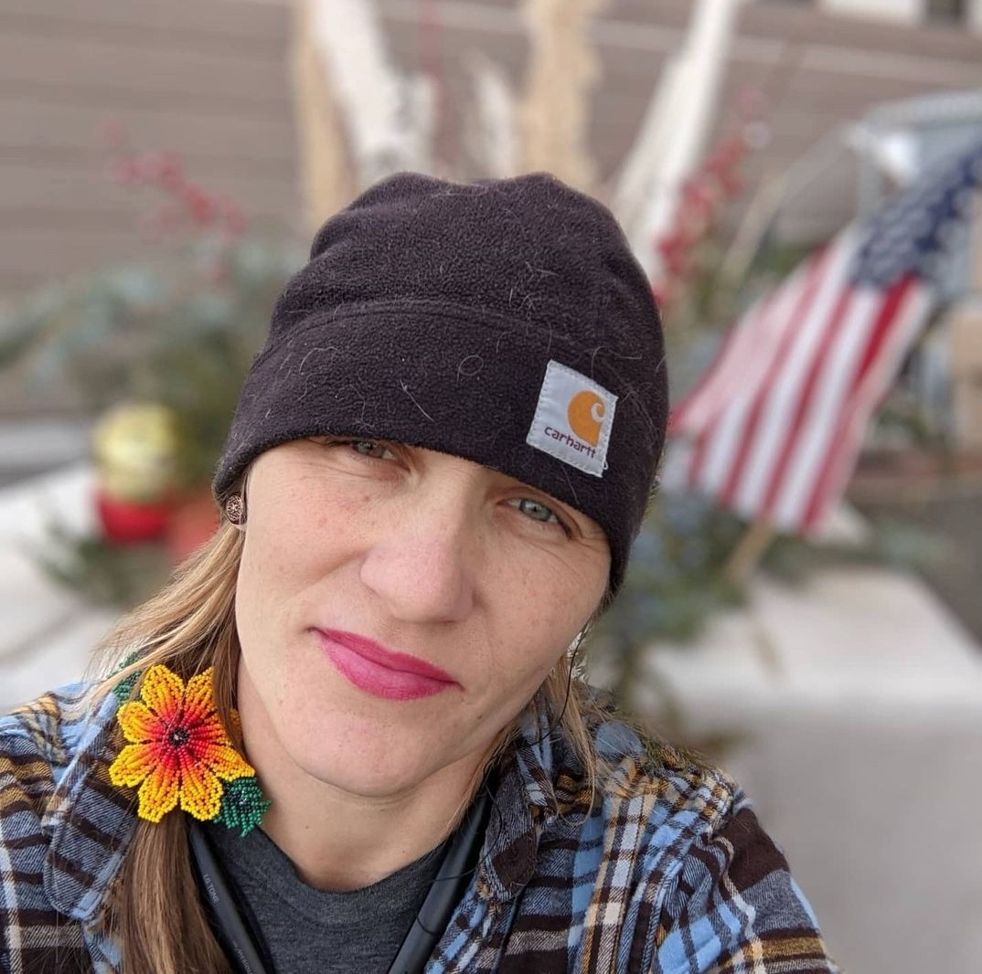Sally Ann is wearing a grey Carharrt hat with a braid and a beaded sunflower clip, a grey shirt, flannel of light blue, yellow and brown. Background is blurred.